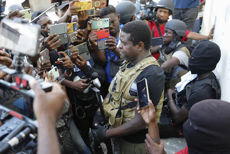 A man in military fatigues is surrounded by people holding microphones and mobile phones.