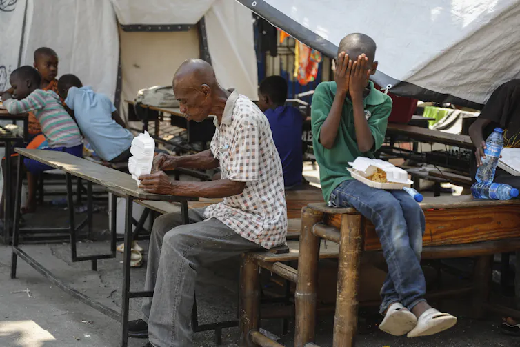 A man sitting in front of a table eats while a child sitting on the table covers his eyes