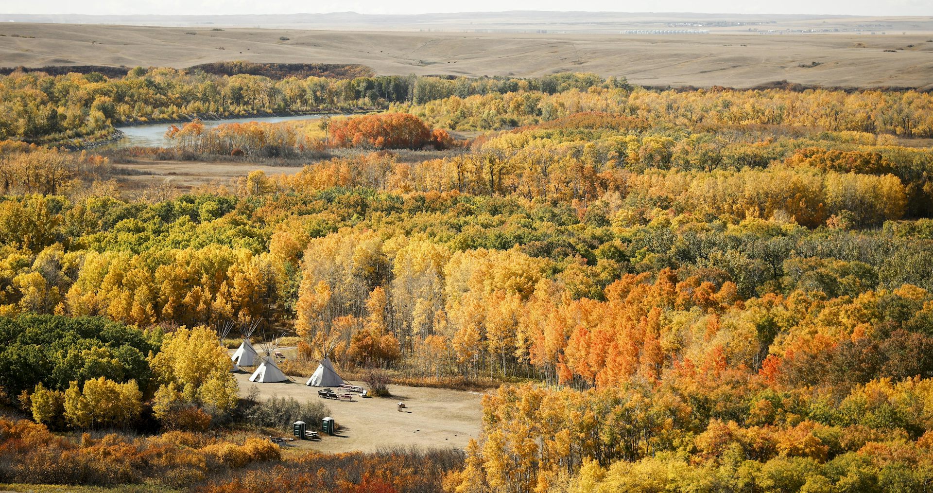 Teepees stand surrounded by fall foliage near a river.