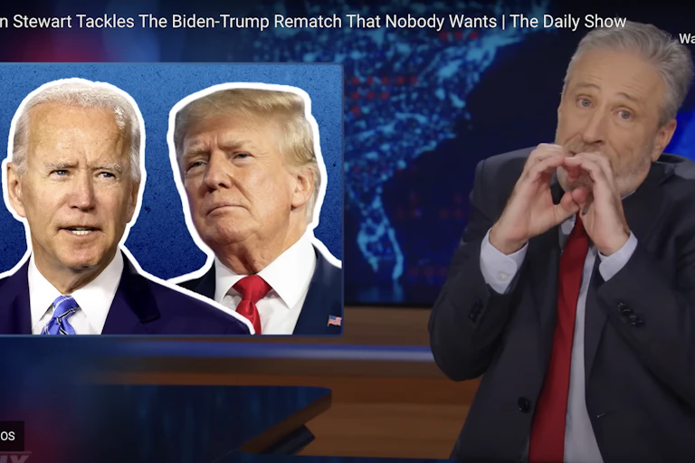 A man making a funny shape with his hands with photos of Donald Trump and Joe Biden on a screen next to him.