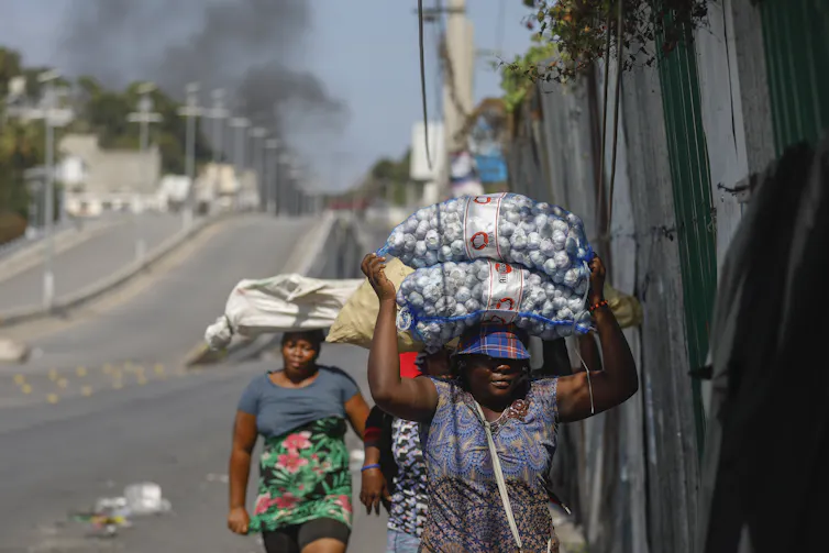 Women carrying filled bags on their heads walk down a street
