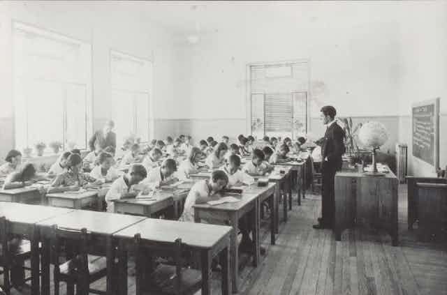 Fotografía en blanco clase con niños y niñas escribiendo ante la mirada de un maestro. y negro de una