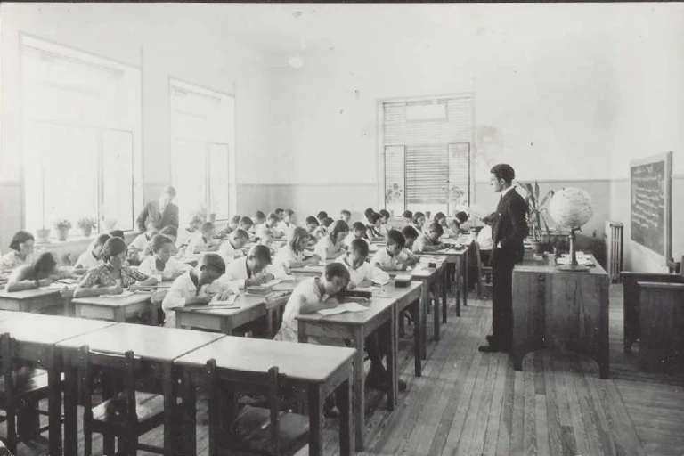 Fotografía en blanco clase con niños y niñas escribiendo ante la mirada de un maestro. y negro de una