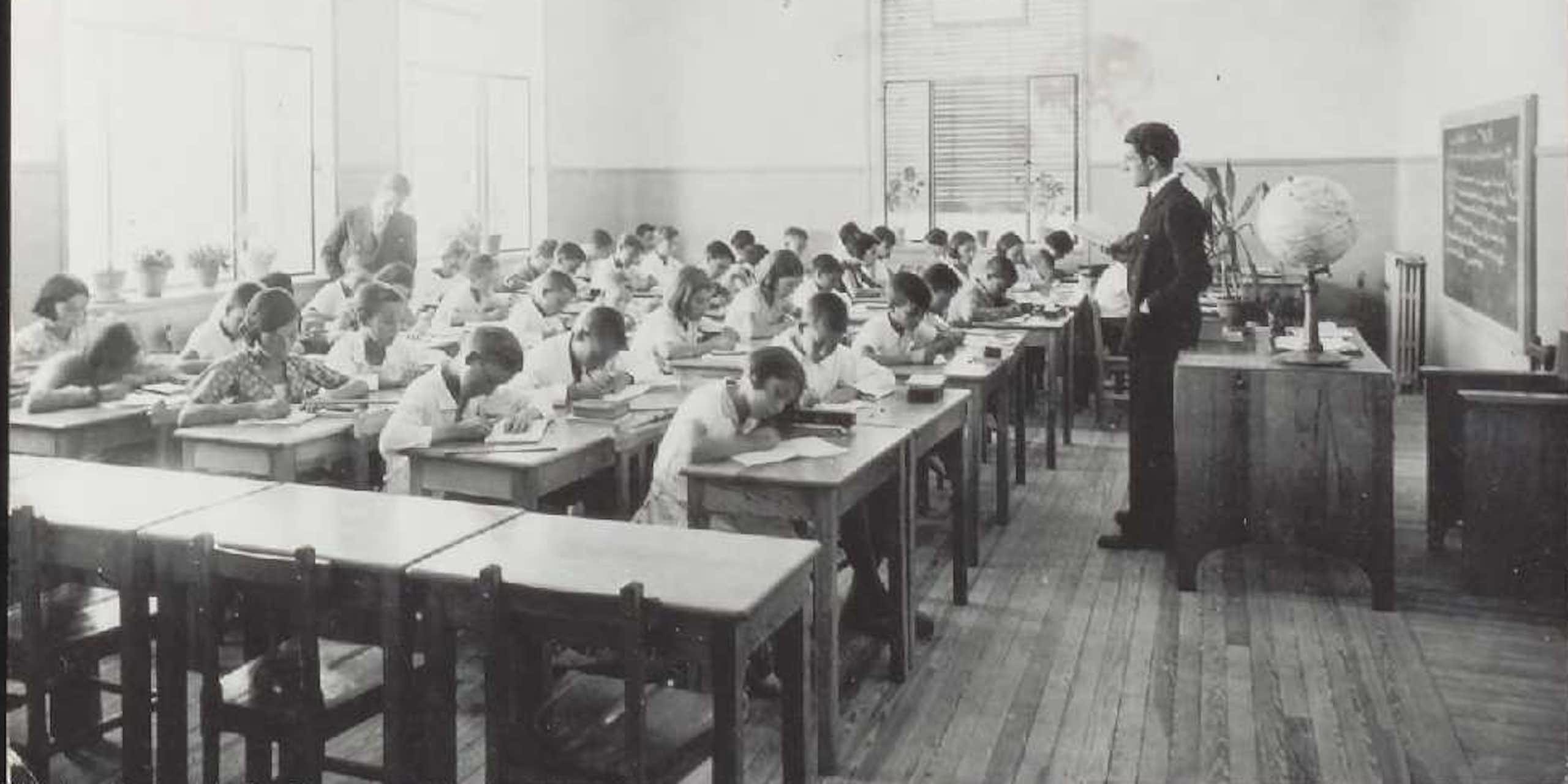 Fotografía en blanco clase con niños y niñas escribiendo ante la mirada de un maestro. y negro de una