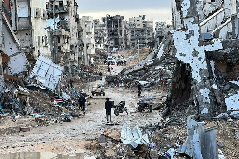 The rubble of destroyed city buildings, with a few people wandering through.
