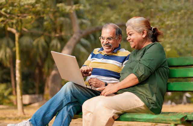 An older man and woman sit on a park bench using a laptop.