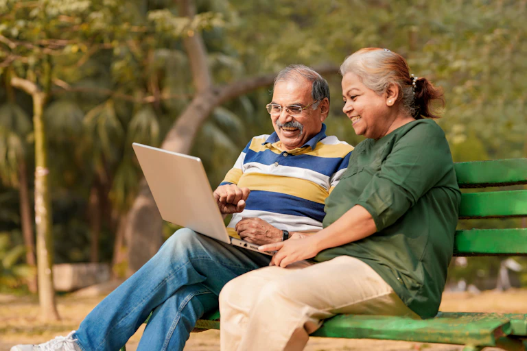 An older man and woman sit on a park bench using a laptop.