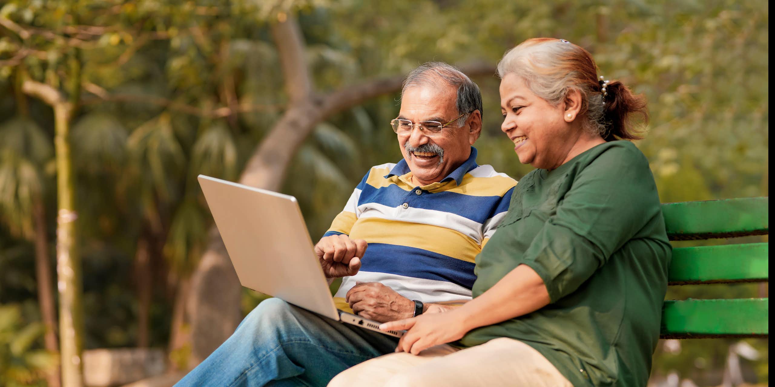 An older man and woman sit on a park bench using a laptop.