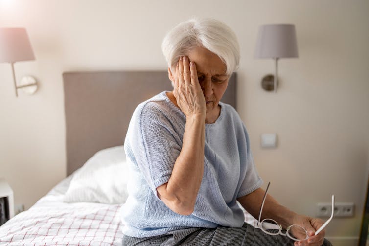 A senior woman sitting on a bed with her hand to her face.