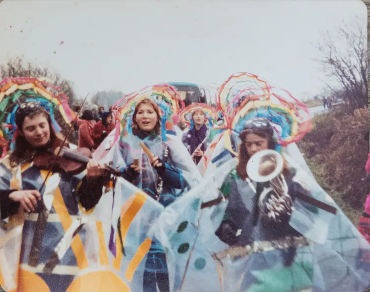 Women protesting wearing rainbow crowns and plastic raincoats. One holds a megaphone.