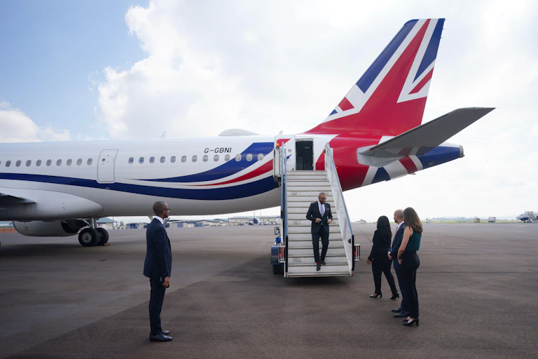 James Cleverly walks off the plane at at Kigali International Airport in Rwanda. He is at the bottom of a flight of stairs off of a large plane with the tail in a UK flag design