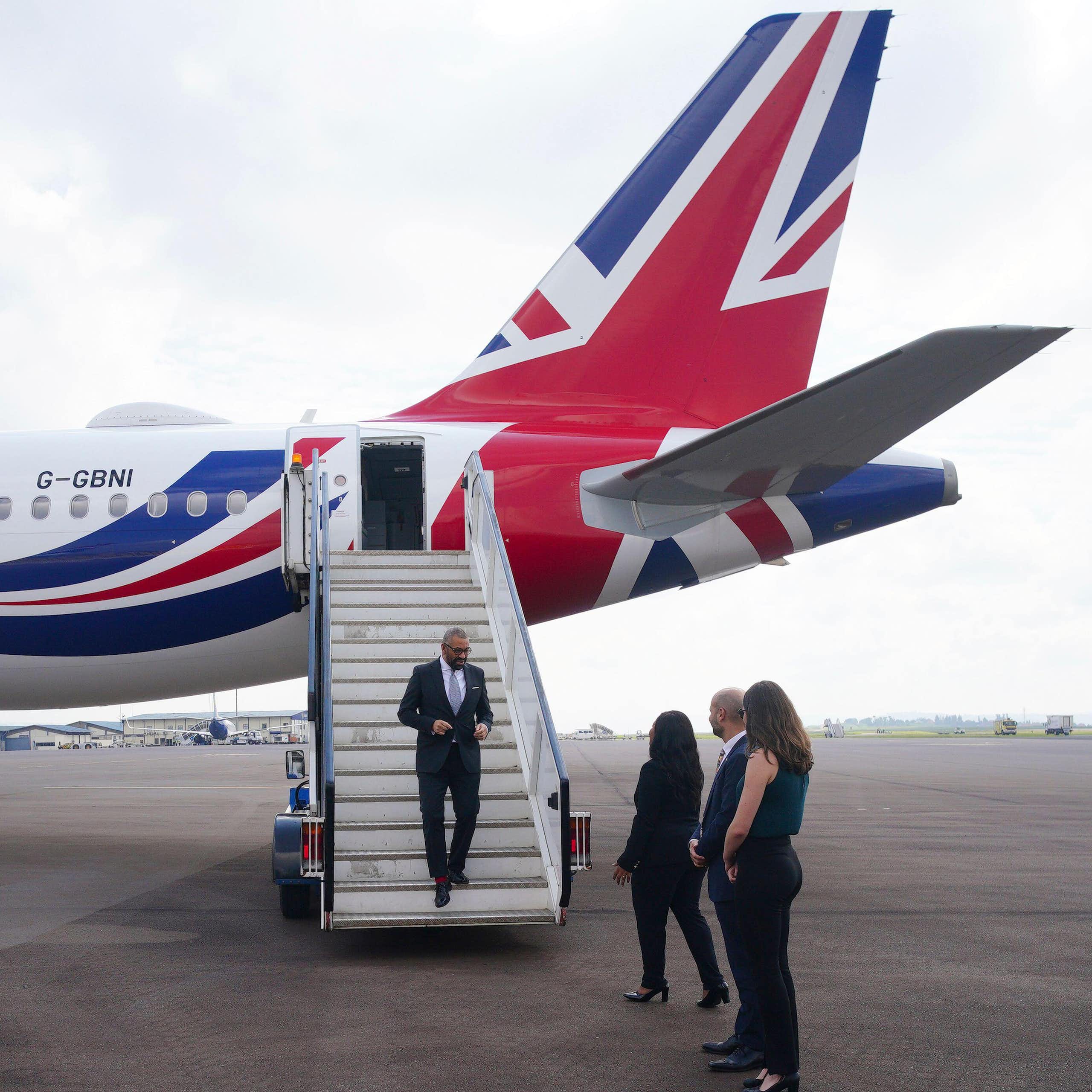 James Cleverly walks off the plane at at Kigali International Airport in Rwanda. He is at the bottom of a flight of stairs off of a large plane with the tail in a UK flag design