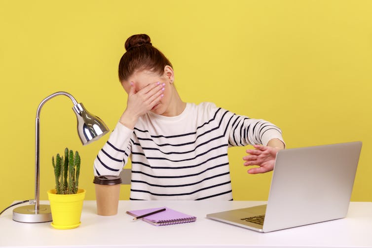 A young woman sitting at a desk in front of a yellow wall covers her eyes with one hand and uses the other to hide her computer screen, suggesting that she doesn't want to see this.
