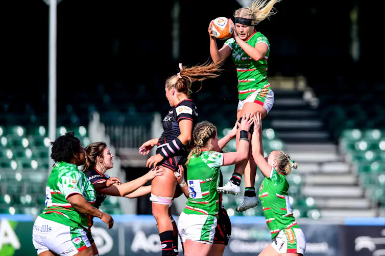 A lineout in a women's rugby game where a player in green is hoisted above the other players.