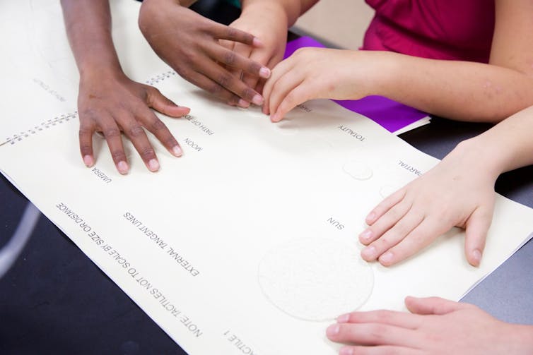 A sheet of paper with raised textures labeled Sun Umbra Moon and Totality with three students touching the textures