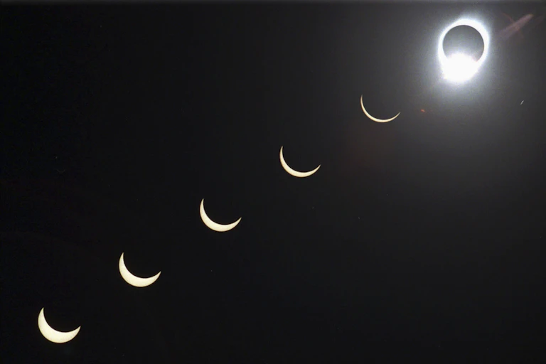 A solar eclipse, with the Moon covering the Sun with just an outline of light visible around it, in the top right, with varying stages of coverage moving from the bottom left to the right, as the sliver of Sun visible gets smaller.