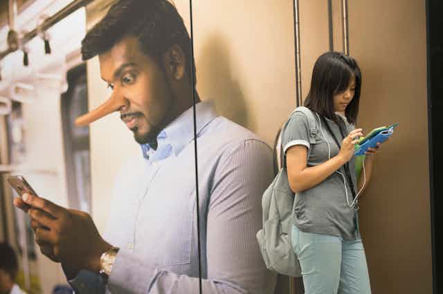 A woman looks at her phone in front of a large poster of a man also looking at his phone with a long Pinocchio nose.