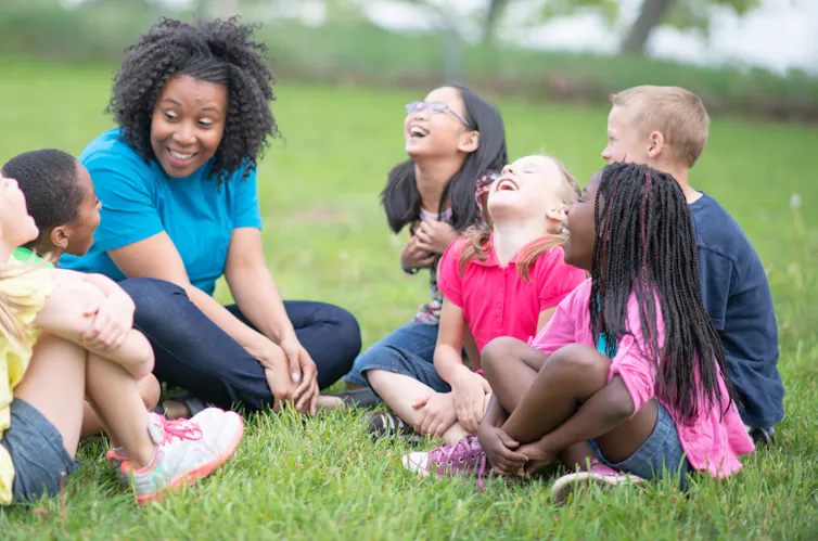 A teacher and several children sit on the grass at a park and laugh.