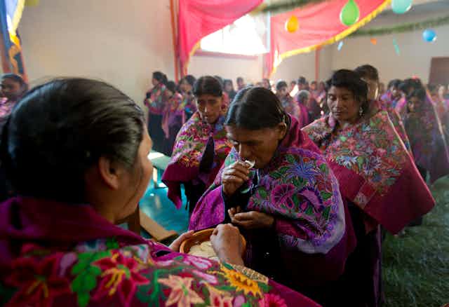Lines of women in richly embroidered red and purple shawls, with a woman in front holding a plate with wafers on it.