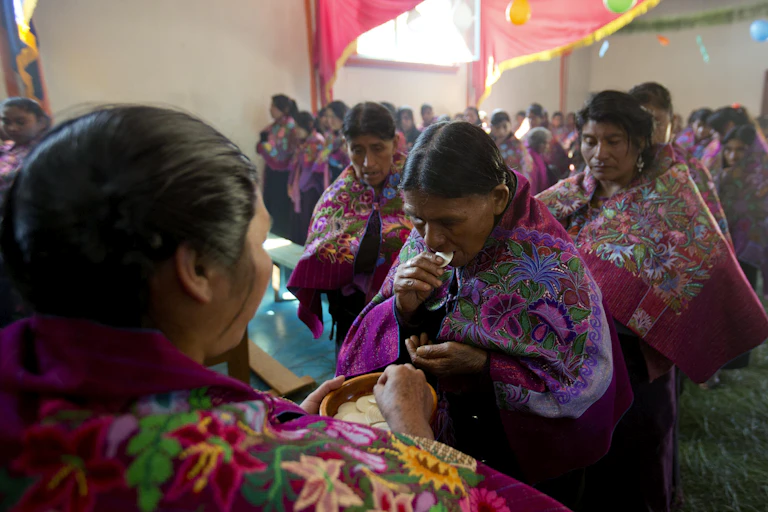 Lines of women in richly embroidered red and purple shawls, with a woman in front holding a plate with wafers on it.