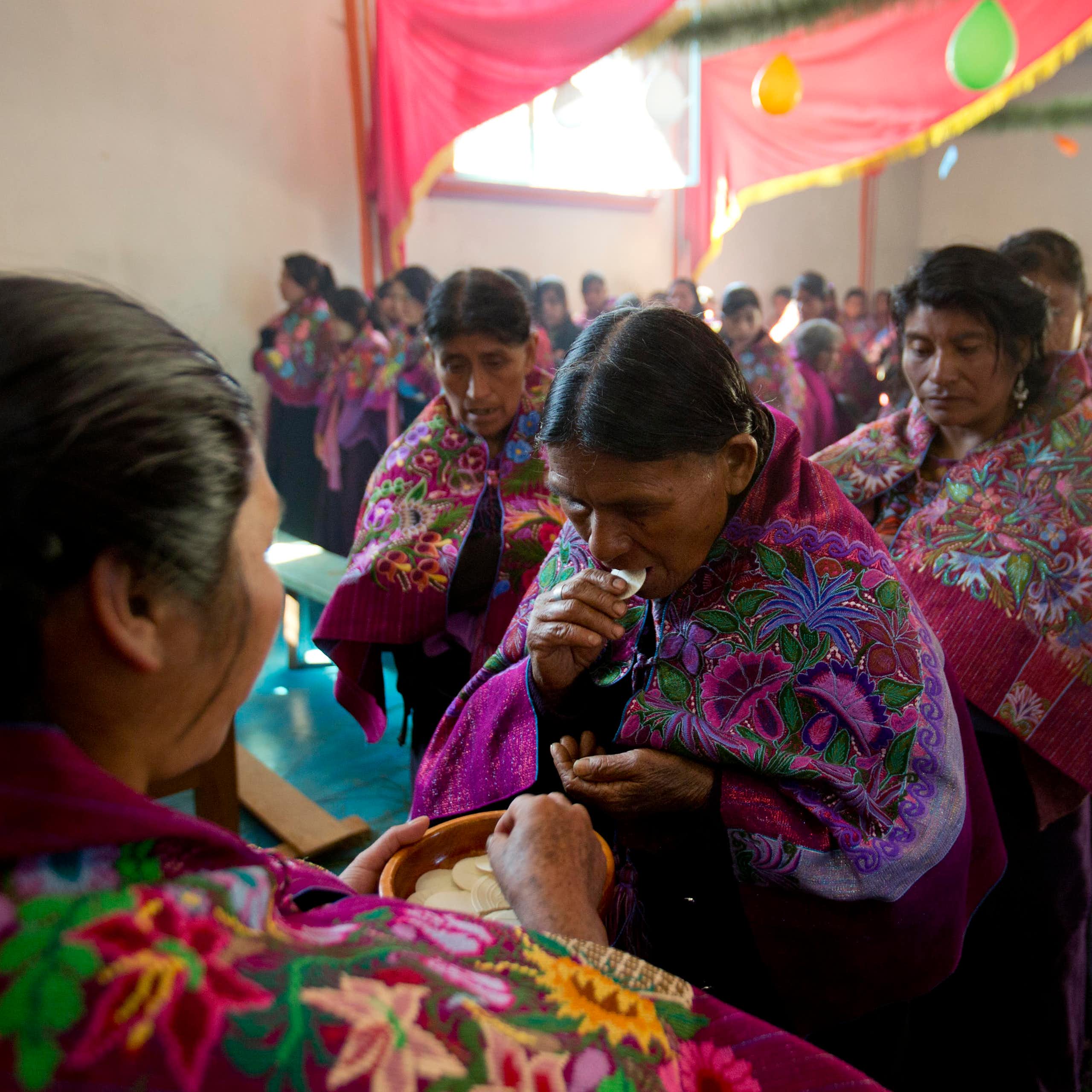 Lines of women in richly embroidered red and purple shawls, with a woman in front holding a plate with wafers on it.