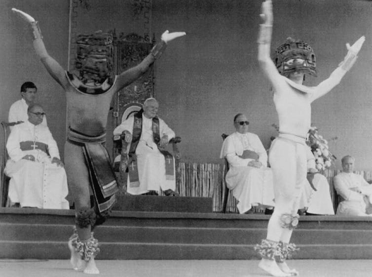 A black and white photo shows several seated men in white watching two men in headdresses dance with their arms raised.