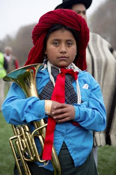 A boy in a red headdress and bright blue shirt stands holding a small brass instrument.