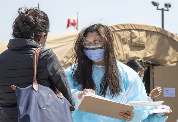 A dark-haired woman with a mask on and wearing medical scrubs talks to another woman with her back to the camera on a sunny day.