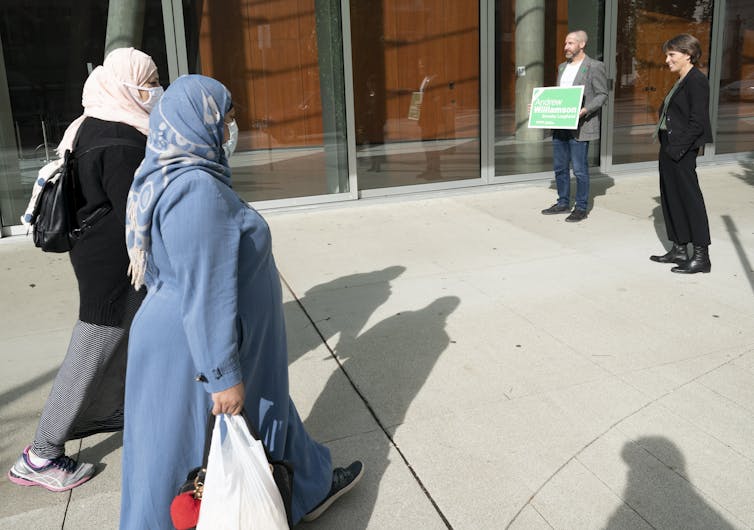 Two women in headscarves carrying shopping bags walk past a politician.