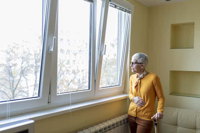 An elderly woman holding on to a cane looks out a window.