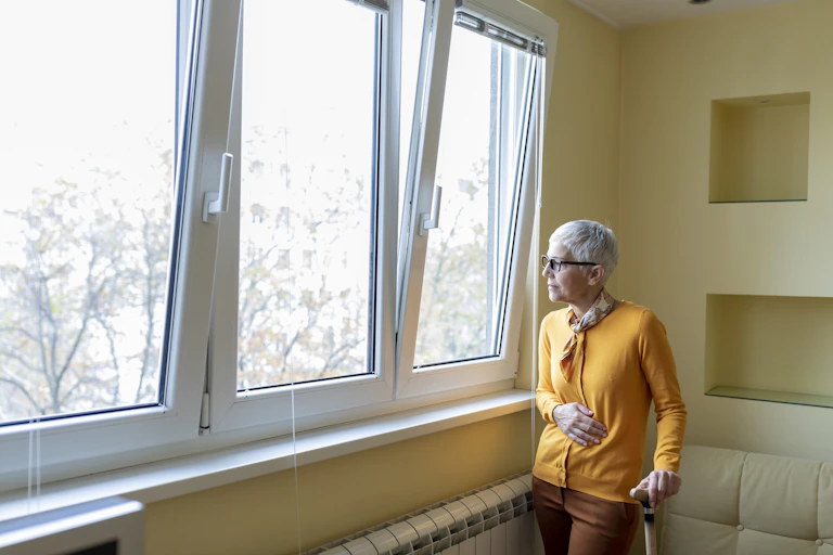 An elderly woman holding on to a cane looks out a window.