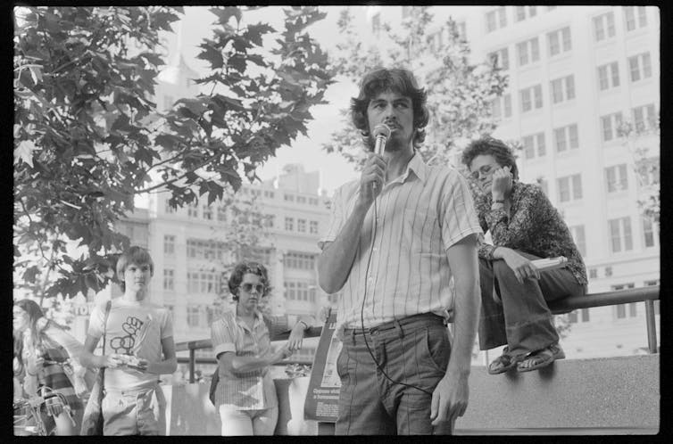 a man talking into a microphone, in a shirt and trousers, with onlookers listening