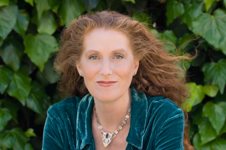 A woman with long red hair smiling in front of a wall of ivy