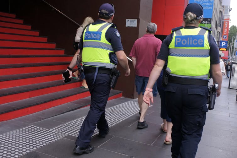 Two people with high-vis vests that have police written on the back walk down a city street.