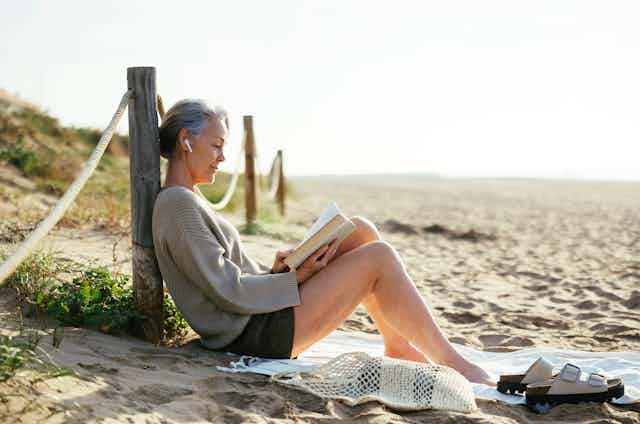 Mature woman wearing wireless in ear headphones reading book at beach 