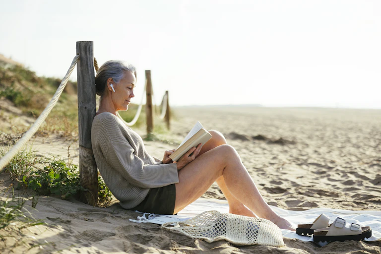 Mature woman wearing wireless in ear headphones reading book at beach