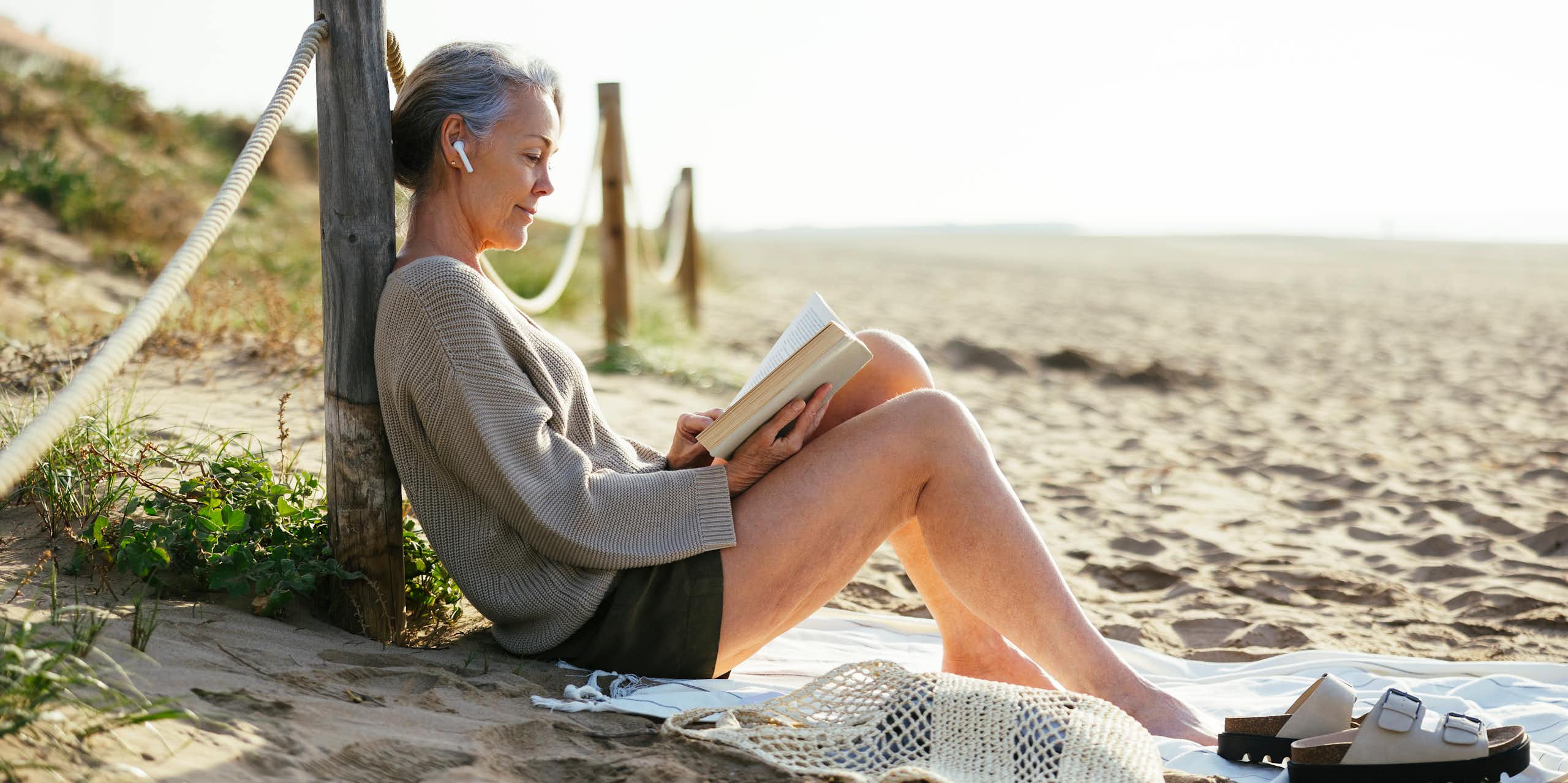 Mature woman wearing wireless in ear headphones reading book at beach 