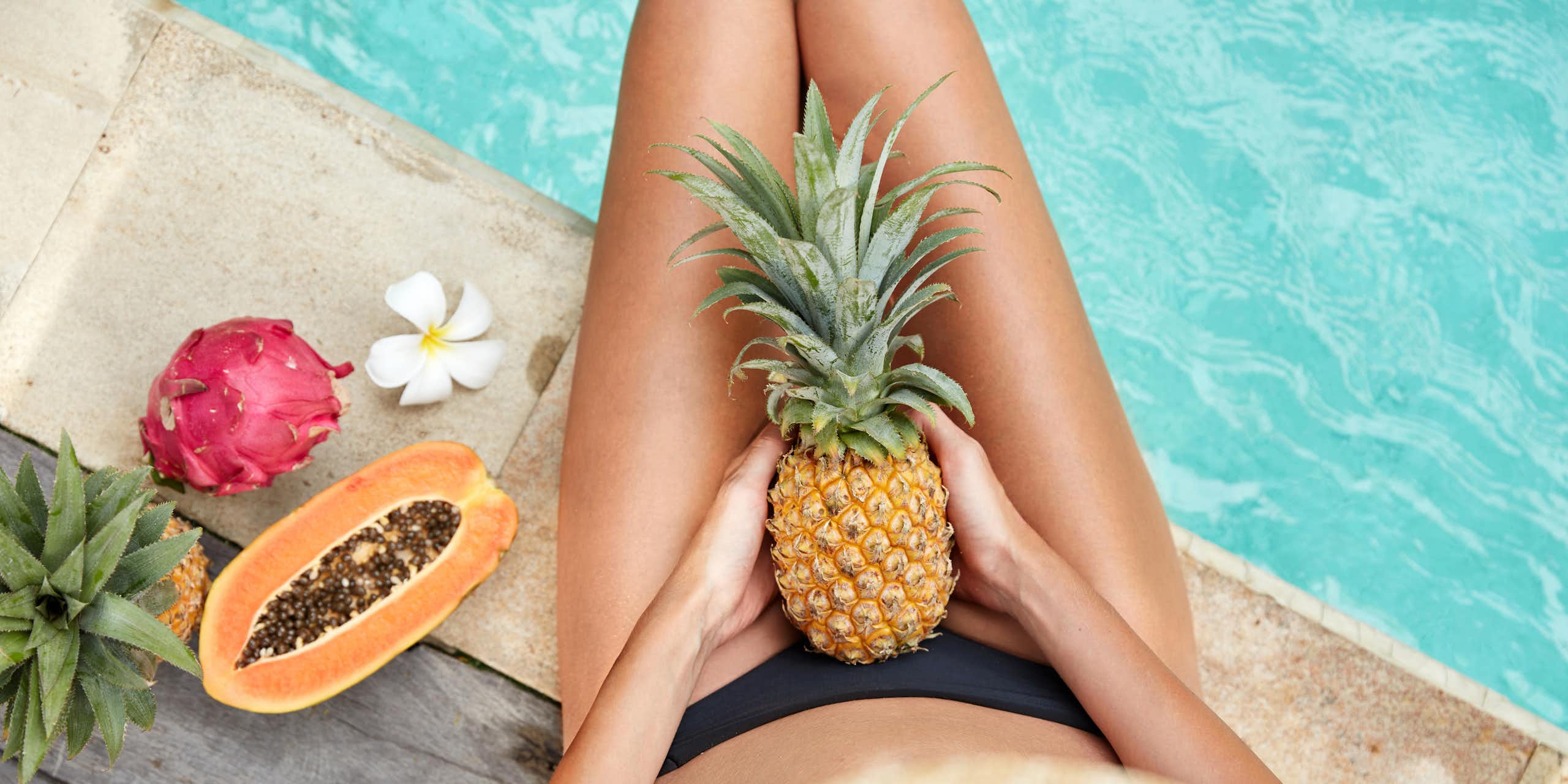 Woman in a bikini, sitting next to a pool, holding a pineapple on her lap