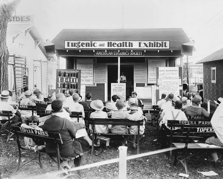 Photographie en noir et blanc. Un public assis, vu de dos, est tourné vers une présentation sur un stand nommé « Eugenic and Health Exhibit »