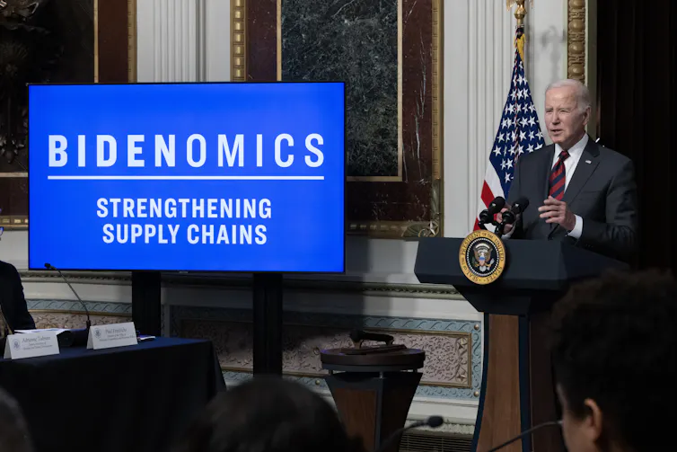 President Biden speaking next to screen showing the word 'Bidenomics'.