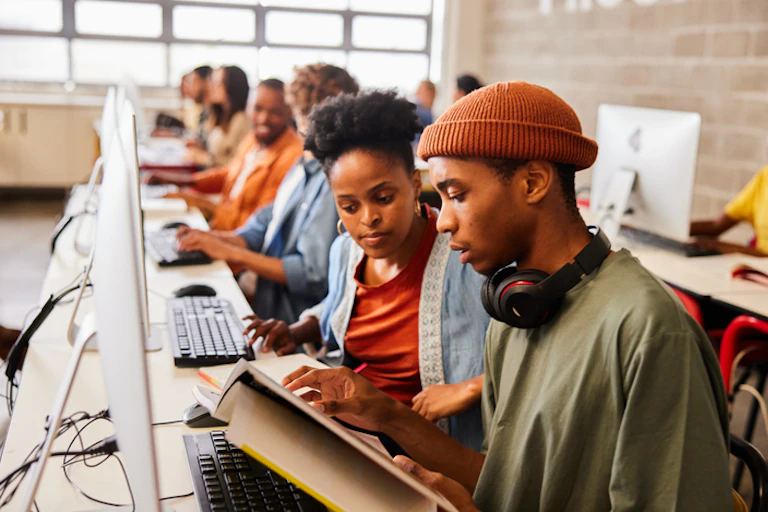 A young woman looks at a book alongside a young man with headphones around his neck. They're both seated at computers.