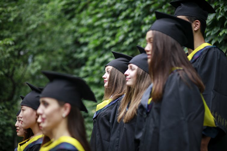 Seven college students wearing black graduation gowns and caps face left. They're wearing yellow stoles.