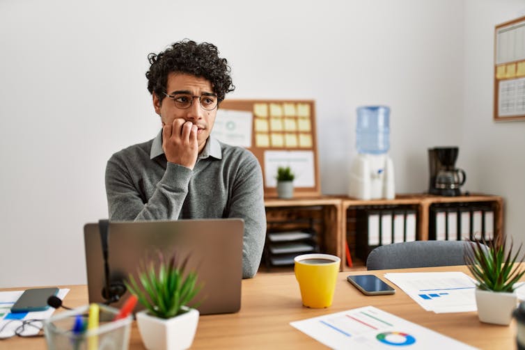 A young man with a nervous look on his face sits at a table in an office while biting hit nails