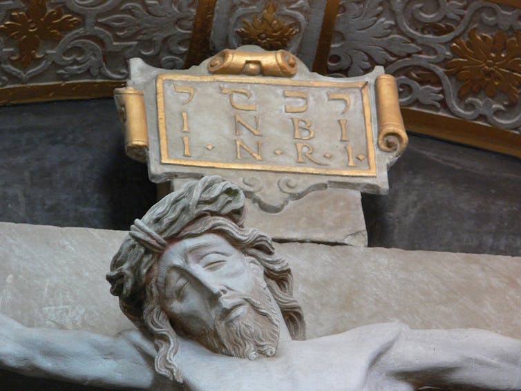 A close-up photo of a pale sculpture of a bearded man's face, looking in pain or tired, with gold letters above.