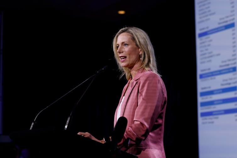 A woman in a pink blazer delivers a speech behind a lectern.