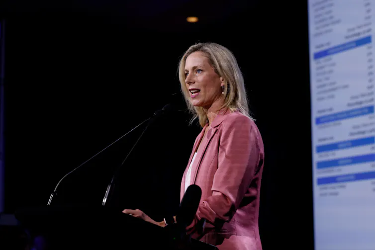 A woman in a pink blazer delivers a speech behind a lectern.