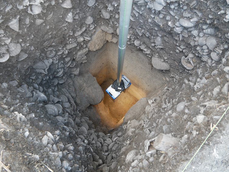 A photo showing a device being lowered into a deep square pit. The dirt walls of the pit are fills with sea shells.