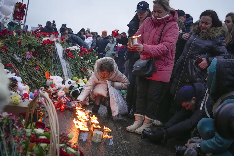 One woman kneels to light a candle while a crowd of other mourners look on.