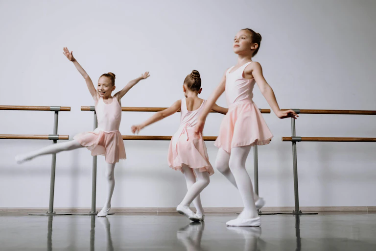 Three young girls in ballet costumes dance next to a barre.