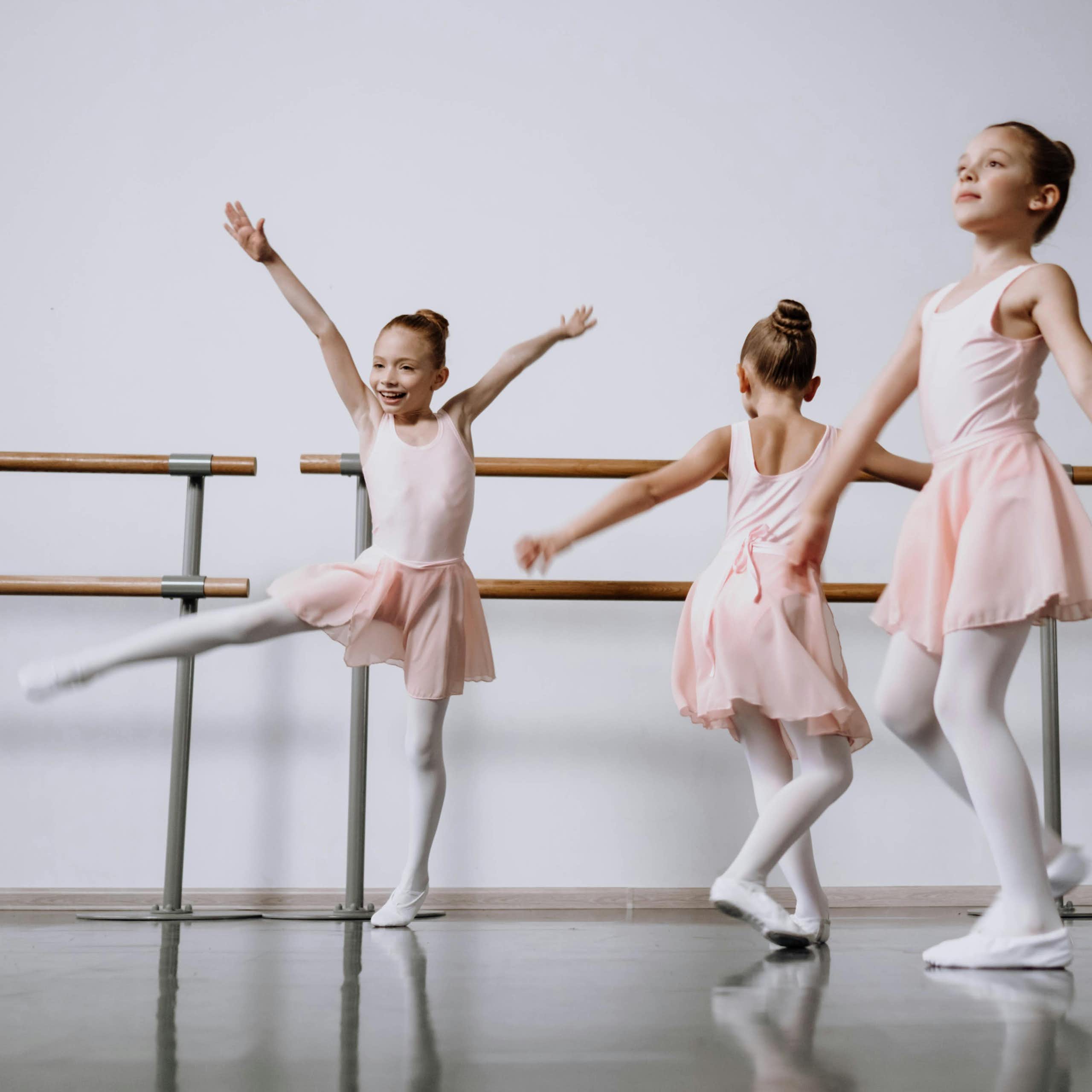Three young girls in ballet costumes dance next to a barre.